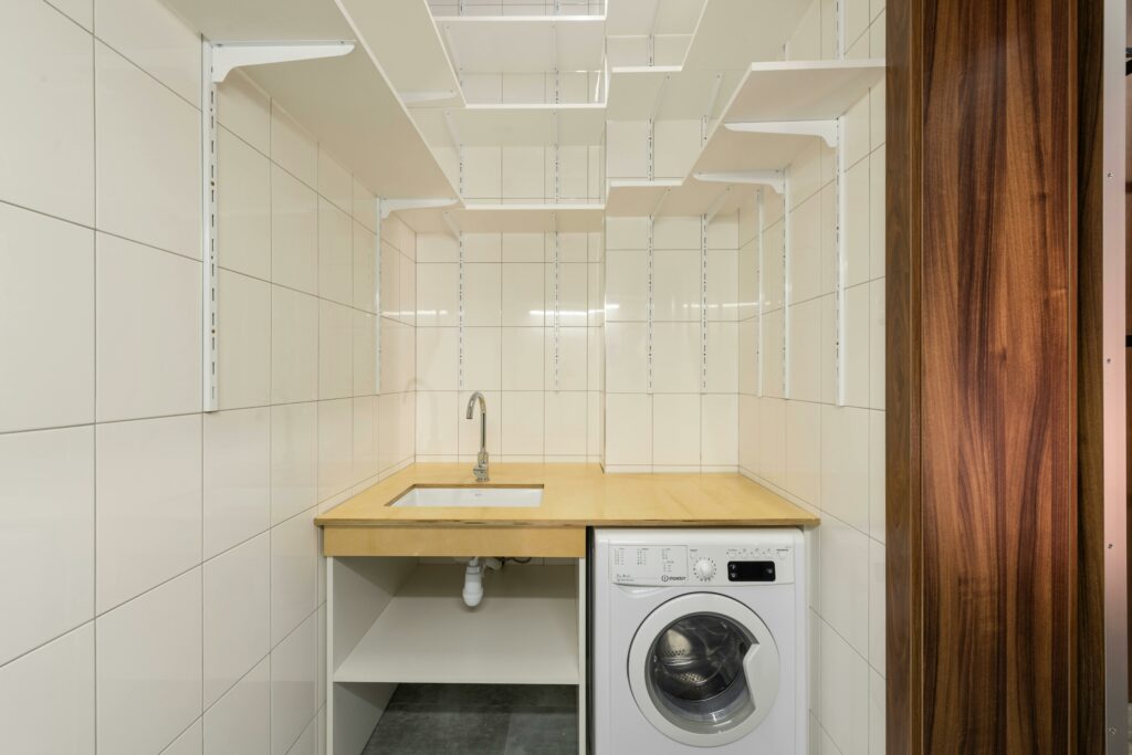 Lavanderia residencial organizada e limpa com azulejos brancos, prateleiras de armazenamento superiores e uma máquina de lavar embutida sob uma bancada de madeira, destacando um design minimalista e funcional.