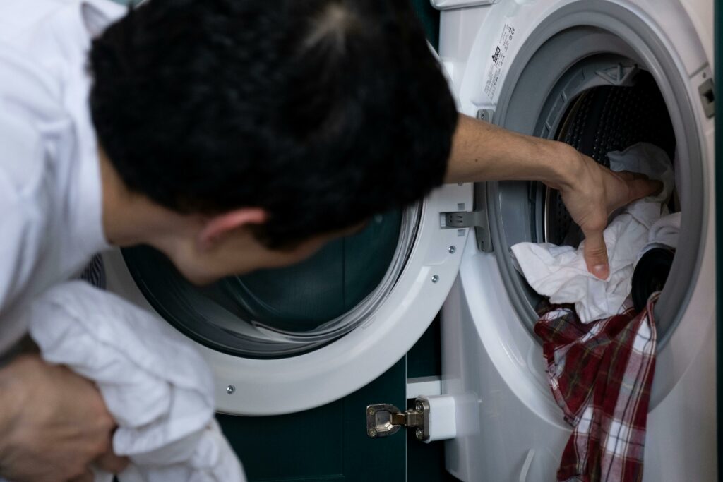 Pessoa carregando ou descarregando uma máquina de lavar frontal, focada na tarefa de manuseio das roupas, em um ambiente que parece ser uma área de serviço de uma casa ou apartamento.