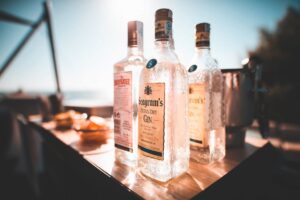 A imagem mostra três garrafas de gin com destaque para marcas conhecidas. As garrafas estão posicionadas sobre uma mesa, possivelmente num ambiente externo, dada a luz do sol que ilumina a cena com tons dourados e quentes. A primeira garrafa à esquerda é da marca Beefeater London Dry Gin, reconhecível pelo seu rótulo rosa e logo distinto. Ao centro, há uma garrafa de Seagram's Extra Dry Gin, com seu rótulo característico que remete a um design clássico. A terceira garrafa, à direita, é de Gordon's London Dry Gin, com um rótulo que combina cores e tipografia tradicionais. As garrafas estão cobertas de condensação, sugerindo que estão geladas, o que é ideal para servir um gin fresco e revigorante. O fundo desfocado indica um ambiente de lazer e relaxamento, talvez uma varanda ou um deck com vista para uma paisagem, sugerindo um momento de desfrute e descontração.