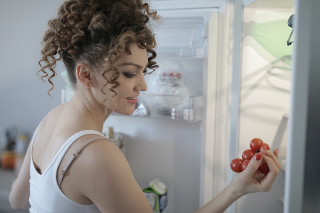 Mulher branca com blusa branca cabelos cacheados segurando tomates em frente a uma geladeira frost free aberta.