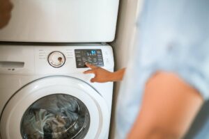 Uma vista de cima de uma máquina de lavar moderna. Uma mão masculina está ajustando as configurações no painel digital da máquina. Podemos identificar o logo "LG" e alguns botões e funções no painel, como "Quick Wash" e "Start/Pause".