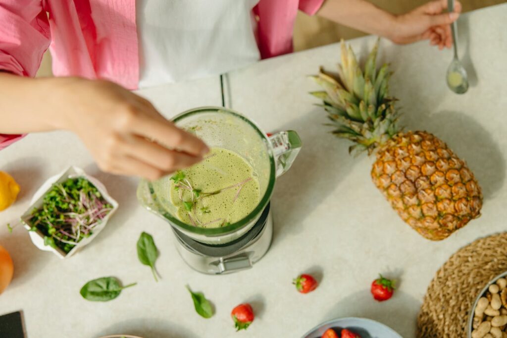 Uma pessoa finalizando a preparação de um suco verde no liquidificador, com ingredientes como abacaxi e espinafre ao redor, sugerindo um estilo de vida saudável.